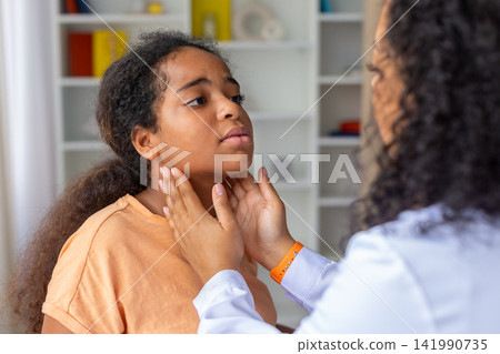 Black female doctor examining young girls neck and throat during pediatric checkup Black female doctor examining young girls neck and throat during pediatric checkup 141990735