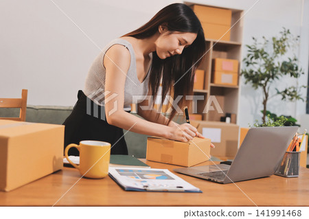 Startup SME small business entrepreneur of freelance Asian woman wearing apron using laptop and box to receive and review orders online to prepare to pack sell to customers, online sme business ideas. Startup SME small business entrepreneur of freelance Asian woman wearing apron using laptop and box to receive and review orders online to prepare to pack sell to customers, online sme business ideas. 141991468