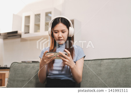 Beautiful young asian woman sitting on sofa and holding joystick playing online video game in living room at house Beautiful young asian woman sitting on sofa and holding joystick playing online video game in living room at house 141991511