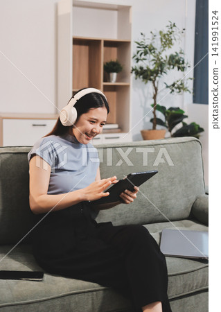 Freelance Asian woman holding mobile phone typing on laptop and working from home. Happy young girl sitting on sofa in living room. Freelance Asian woman holding mobile phone typing on laptop and working from home. Happy young girl sitting on sofa in living room. 141991524