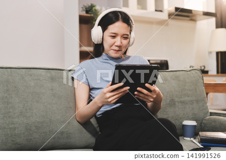 Freelance Asian woman holding mobile phone typing on laptop and working from home. Happy young girl sitting on sofa in living room. Freelance Asian woman holding mobile phone typing on laptop and working from home. Happy young girl sitting on sofa in living room. 141991526