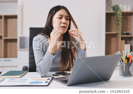 Young Asian woman sits at office desk rubbings her eyes and temples feeling pain. Stress. And exhaustion from long hours working on the computer Young Asian woman sits at office desk rubbings her eyes and temples feeling pain. Stress. And exhaustion from long hours working on the computer 141991545