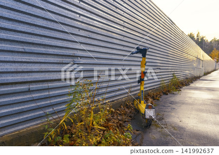Yellow electric scooter parked beside corrugated metal fence on wet sidewalk at dusk Yellow electric scooter parked beside corrugated metal fence on wet sidewalk at dusk 141992637