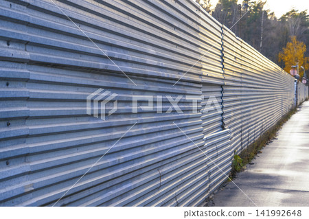 Corrugated metal construction fence stretching along wet sidewalk at dusk with reflection and distant trees Corrugated metal construction fence stretching along wet sidewalk at dusk with reflection and distant trees 141992648