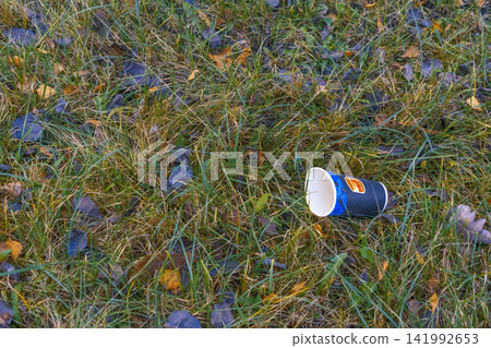 Discarded paper cup lying in grassy field with autumn leaves, showing outdoor litter and seasonal neglect Discarded paper cup lying in grassy field with autumn leaves, showing outdoor litter and seasonal neglect 141992653