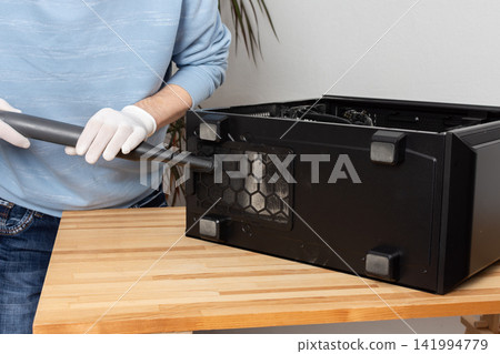 A 53-year-old man uses a vacuum to clean dust from a computer system unit on a wooden table in a home office. The setting is tidy and organized A 53-year-old man uses a vacuum to clean dust from a computer system unit on a wooden table in a home office. The setting is tidy and organized 141994779