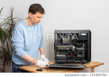 A 53-year-old man removes dust from a computer system unit on a wooden table. He is focused on cleaning the inside of the computer in a well-lit room 141994780