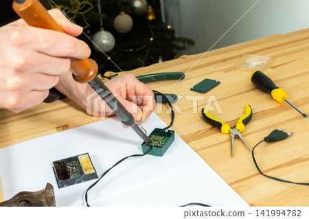 A man works on fixing a broken Christmas tree garland by soldering wires together. He uses tools and parts on a wooden table, focusing on the repair process during the holiday season A man works on fixing a broken Christmas tree garland by soldering wires together. He uses tools and parts on a wooden table, focusing on the repair process during the holiday season 141994782