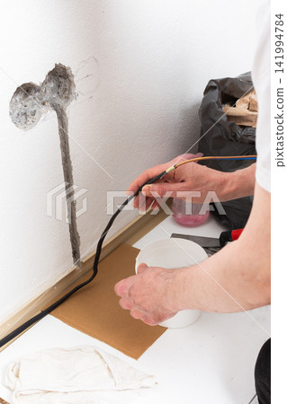 Worker runs new electrical wires into a cutout in a white apartment wall. This task prepares for the installation of an electrical outlet near the baseboard Worker runs new electrical wires into a cutout in a white apartment wall. This task prepares for the installation of an electrical outlet near the baseboard 141994784