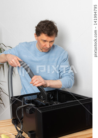 A 53-year-old man uses a vacuum attachment to remove dust from a computer system unit in a home office. The room is well lit and organized for the task A 53-year-old man uses a vacuum attachment to remove dust from a computer system unit in a home office. The room is well lit and organized for the task 141994795
