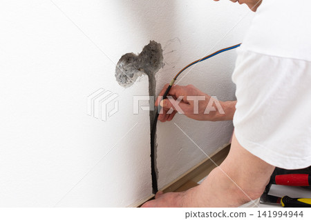 Worker runs new electrical wires into a cutout in a white apartment wall. This task prepares for the installation of an electrical outlet near the baseboard Worker runs new electrical wires into a cutout in a white apartment wall. This task prepares for the installation of an electrical outlet near the baseboard 141994944