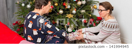 A couple sits near a Christmas tree on Christmas Day. They are exchanging gifts and enjoying the holiday spirit. The room has decorations and a festive atmosphere A couple sits near a Christmas tree on Christmas Day. They are exchanging gifts and enjoying the holiday spirit. The room has decorations and a festive atmosphere 141994945