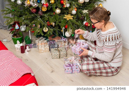 A woman aged 52 sits on the floor next to a decorated Christmas tree. She opens gifts on Christmas Day while enjoying the holiday with family members in the background A woman aged 52 sits on the floor next to a decorated Christmas tree. She opens gifts on Christmas Day while enjoying the holiday with family members in the background 141994946