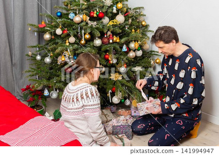 A 52-year-old couple sits on the floor, smiling and opening gifts. The room is decorated with a Christmas tree, colorful ornaments, and presents around them. It is Christmas Day A 52-year-old couple sits on the floor, smiling and opening gifts. The room is decorated with a Christmas tree, colorful ornaments, and presents around them. It is Christmas Day 141994947