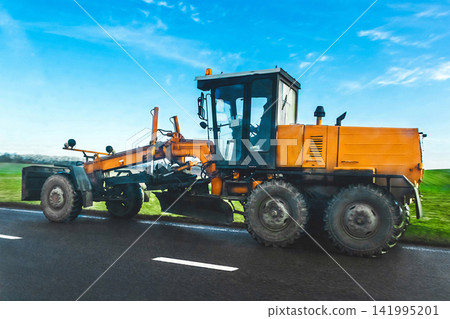The road scraper rides on a asphalt against the green meadow and blue sky. Industrial transport, car The road scraper rides on a asphalt against the green meadow and blue sky. Industrial transport, car 141995201