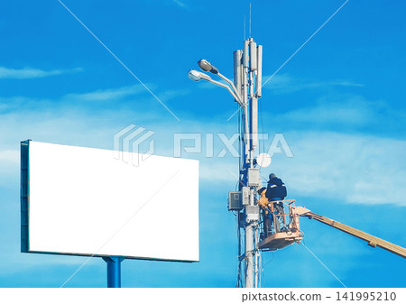 A mock-up large white billboard form and two construction workers on a machine crane repair a lighting pole against a blue sky A mock-up large white billboard form and two construction workers on a machine crane repair a lighting pole against a blue sky 141995210