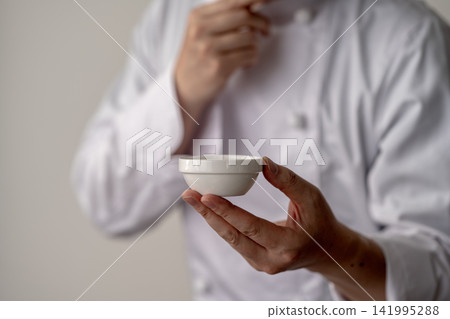 The chef is tasting the food in a small white bowl. 141995288