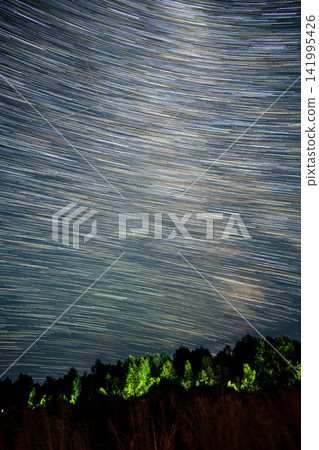 Starry night sky filled with streaking lights. Distant trees glow softly in green. Dark horizon contrasts bright celestial trails. Nighttime capture of motion in the heavens Starry night sky filled with streaking lights. Distant trees glow softly in green. Dark horizon contrasts bright celestial trails. Nighttime capture of motion in the heavens 141995426