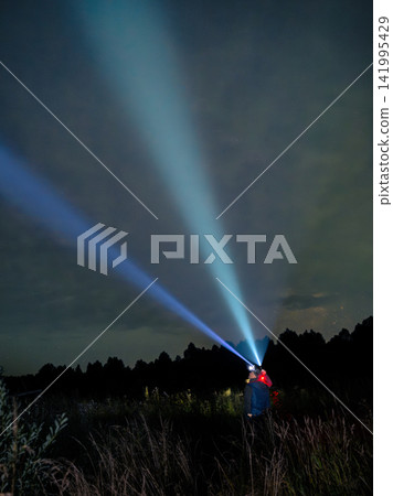 Night sky illuminated by powerful beams from handheld light. Person stands in dark field, casting bright rays upward. Distant trees and grasses frame the scene beneath starry sky Night sky illuminated by powerful beams from handheld light. Person stands in dark field, casting bright rays upward. Distant trees and grasses frame the scene beneath starry sky 141995429