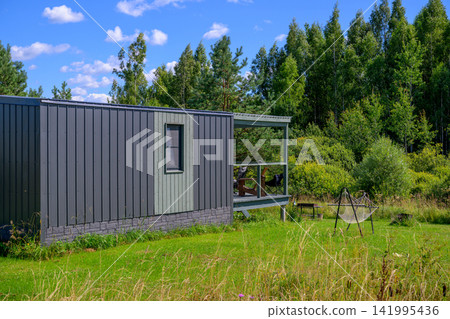 Modern container-style cabin sits in grassy field. Blue sky with scattered white clouds above. Green trees form dense background foliage. Open doorway reveals interior space Modern container-style cabin sits in grassy field. Blue sky with scattered white clouds above. Green trees form dense background foliage. Open doorway reveals interior space 141995436
