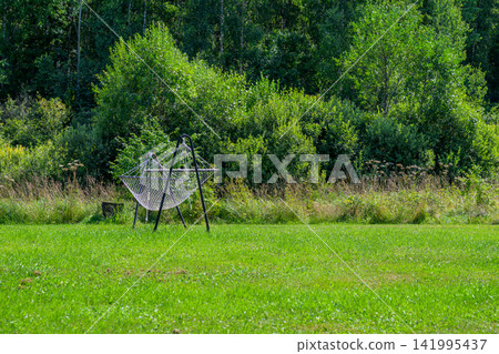Empty swing rests in sunlit green field. Lush trees frame the background. Vines grow along the swing's frame. Sunlight dapples the grassy lawn. Serenity surrounds the quiet scene Empty swing rests in sunlit green field. Lush trees frame the background. Vines grow along the swing's frame. Sunlight dapples the grassy lawn. Serenity surrounds the quiet scene 141995437