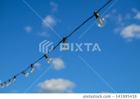 Clear glass bulbs hang from a wire against a bright blue sky. White clouds float gently in the background. Each bulb is simple, unlit, and slightly frosted Clear glass bulbs hang from a wire against a bright blue sky. White clouds float gently in the background. Each bulb is simple, unlit, and slightly frosted 141995438
