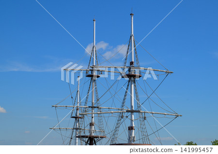 Three tall ship masts stand against a clear blue sky. Metal rigging stretches between sturdy wooden spars. The masts rise sharply, showing maritime engineering Three tall ship masts stand against a clear blue sky. Metal rigging stretches between sturdy wooden spars. The masts rise sharply, showing maritime engineering 141995457