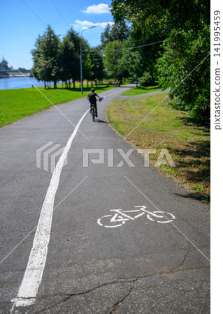 Person rides bicycle on paved path. White line marks bike lane clearly. Lush green trees line both sides. Sky is blue with scattered clouds. Path curves gently through park Person rides bicycle on paved path. White line marks bike lane clearly. Lush green trees line both sides. Sky is blue with scattered clouds. Path curves gently through park 141995459