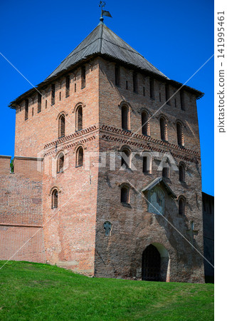 Ancient brick tower stands tall under clear blue sky. Green grass surrounds the stone structure. Weathered walls show signs of age and history. Arched doorway leads into the tower's interior 141995461