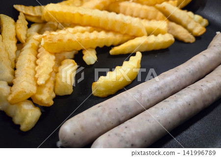 Crinkle-cut French fries served alongside two uncooked sausages on a black frying pan, showcasing a simple meal preparation setting Crinkle-cut French fries served alongside two uncooked sausages on a black frying pan, showcasing a simple meal preparation setting 141996789