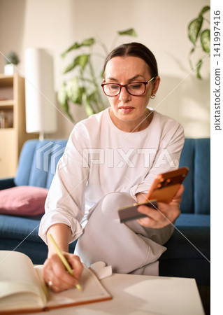 Mature woman confirming online transfer while taking notes at home Mature woman confirming online transfer while taking notes at home 141997416