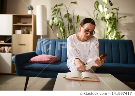Mature woman recording expenses while using smartphone for money transfer Mature woman recording expenses while using smartphone for money transfer 141997417