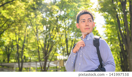 A young man with a backpack on looks up at the sky in the dappled sunlight filtering through the trees. A young man with a backpack on looks up at the sky in the dappled sunlight filtering through the trees. 141997538