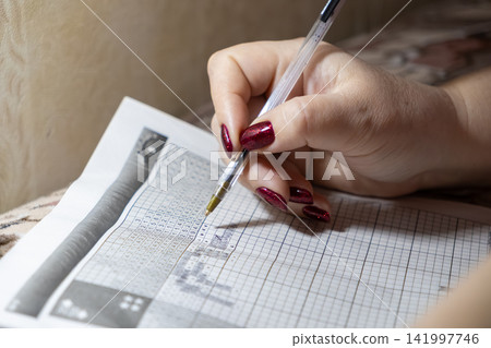 A person with vibrant red nails uses a ballpoint pen to solve a challenging nonogram logic puzzle on paper, illustrating deep mental focus and analytical problem solving skills A person with vibrant red nails uses a ballpoint pen to solve a challenging nonogram logic puzzle on paper, illustrating deep mental focus and analytical problem solving skills 141997746