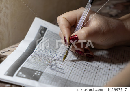 A person with red nails uses a clear ballpoint pen to solve a challenging logic puzzle on grid paper, showcasing focused cognitive activity and a leisurely hobby at home today A person with red nails uses a clear ballpoint pen to solve a challenging logic puzzle on grid paper, showcasing focused cognitive activity and a leisurely hobby at home today 141997747