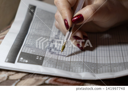 A person with red glittery manicured nails solves a Japanese nonogram logic puzzle using a ballpoint pen to represent deep concentration, cognitive development and mindful leisure A person with red glittery manicured nails solves a Japanese nonogram logic puzzle using a ballpoint pen to represent deep concentration, cognitive development and mindful leisure 141997748