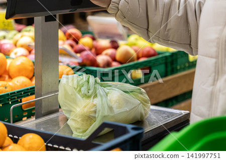 A person carefully weighs a plastic bag of fresh produce on a digital supermarket scale, highlighting healthy shopping habits and daily food consumption in a vibrant grocery store A person carefully weighs a plastic bag of fresh produce on a digital supermarket scale, highlighting healthy shopping habits and daily food consumption in a vibrant grocery store 141997751