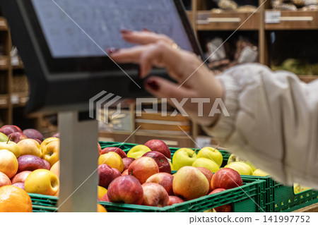 Customer touching a digital scale display in a supermarket to weigh fresh apples, representing modern retail technology, grocery shopping automation, and food industry self-service 141997752