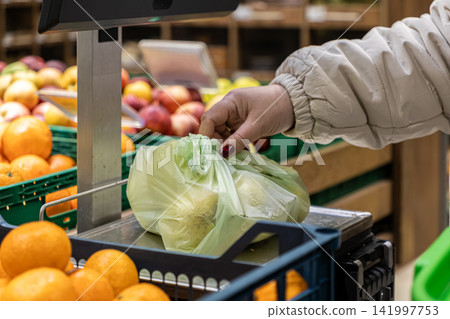 Person weighing a plastic produce bag of fresh fruit on a store scale, highlighting the routine shopping process in a grocery market setting for healthy food and retail convenience Person weighing a plastic produce bag of fresh fruit on a store scale, highlighting the routine shopping process in a grocery market setting for healthy food and retail convenience 141997753