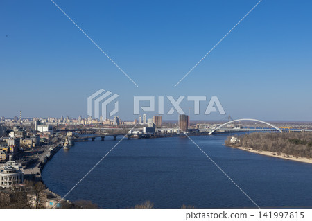 A panoramic view of the Kyiv cityscape featuring the Dnieper river and landmark arched bridge, capturing urban infrastructure on a clear sunny day with a vast blue sky overhead A panoramic view of the Kyiv cityscape featuring the Dnieper river and landmark arched bridge, capturing urban infrastructure on a clear sunny day with a vast blue sky overhead 141997815
