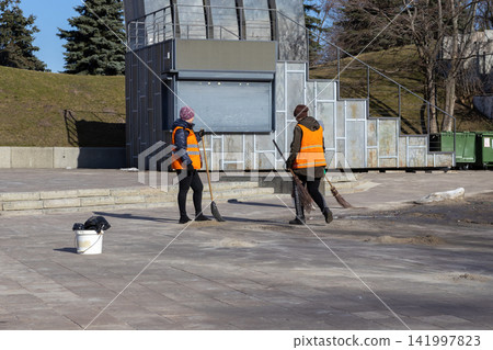 Two municipal workers in orange safety vests sweep a concrete plaza with straw brooms, illustrating essential public maintenance efforts and urban hygiene tasks in city light Two municipal workers in orange safety vests sweep a concrete plaza with straw brooms, illustrating essential public maintenance efforts and urban hygiene tasks in city light 141997823