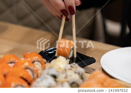Person with red manicured nails holding a salmon sushi roll with chopsticks, dipping it into soy sauce at a restaurant table to highlight a Japanese fine dining culinary experience Person with red manicured nails holding a salmon sushi roll with chopsticks, dipping it into soy sauce at a restaurant table to highlight a Japanese fine dining culinary experience 141997832