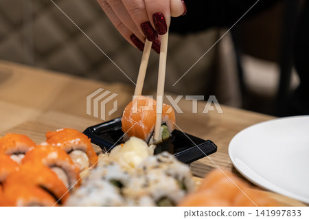 A woman with vibrant red glitter nails holds a fresh salmon sushi roll with wooden chopsticks to dip in soy sauce at a restaurant, showcasing authentic Japanese gourmet food culture A woman with vibrant red glitter nails holds a fresh salmon sushi roll with wooden chopsticks to dip in soy sauce at a restaurant, showcasing authentic Japanese gourmet food culture 141997833