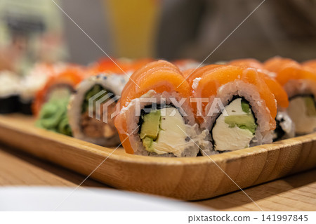 Fresh salmon sushi rolls with cream cheese and avocado arranged on a wooden serving tray, evoking a premium dining experience at a restaurant or a relaxed social gathering meal Fresh salmon sushi rolls with cream cheese and avocado arranged on a wooden serving tray, evoking a premium dining experience at a restaurant or a relaxed social gathering meal 141997845