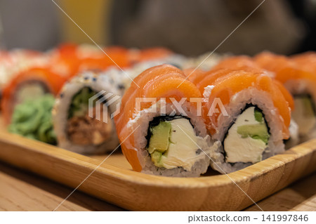 Fresh salmon sushi rolls filled with avocado and cream cheese on a bamboo serving tray, creating an appealing presentation for Japanese cuisine dining in a modern restaurant setup Fresh salmon sushi rolls filled with avocado and cream cheese on a bamboo serving tray, creating an appealing presentation for Japanese cuisine dining in a modern restaurant setup 141997846