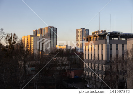 Modern city skyline with skyscrapers catching warm golden hour sunlight above bare winter trees, representing urban growth and architectural contrast in a quiet neighborhood area Modern city skyline with skyscrapers catching warm golden hour sunlight above bare winter trees, representing urban growth and architectural contrast in a quiet neighborhood area 141997873