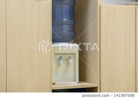 Water cooler dispenser with a large blue plastic bottle inside a light wooden kitchen cabinet unit, providing clean hydration for staff in a professional modern office environment 141997886