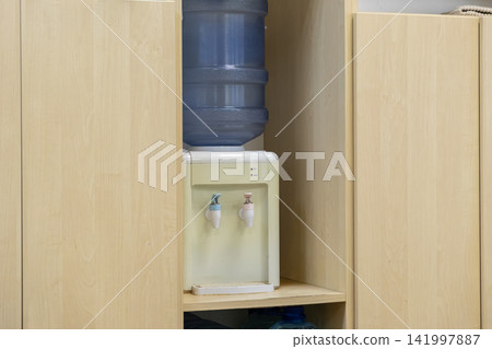 Water dispenser with a blue plastic jug sitting in a wooden kitchen cabinet niche, highlighting a clean and functional refreshment station for an office or a modern residential home Water dispenser with a blue plastic jug sitting in a wooden kitchen cabinet niche, highlighting a clean and functional refreshment station for an office or a modern residential home 141997887
