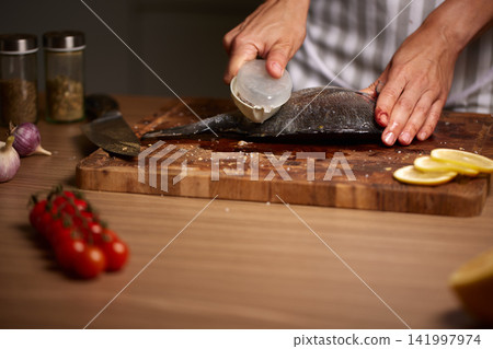 Preparing fresh dorado fish for cooking, removing scales with a specialized tool on a wooden cutting board with garlic and lemon Preparing fresh dorado fish for cooking, removing scales with a specialized tool on a wooden cutting board with garlic and lemon 141997974