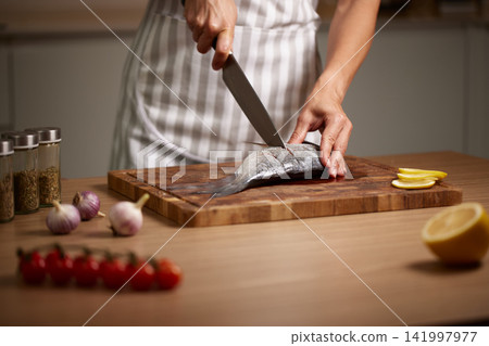 Hands of a cook scoring fresh sea bream fish on a wooden cutting board with a knife, preparing for a healthy seafood meal Hands of a cook scoring fresh sea bream fish on a wooden cutting board with a knife, preparing for a healthy seafood meal 141997977